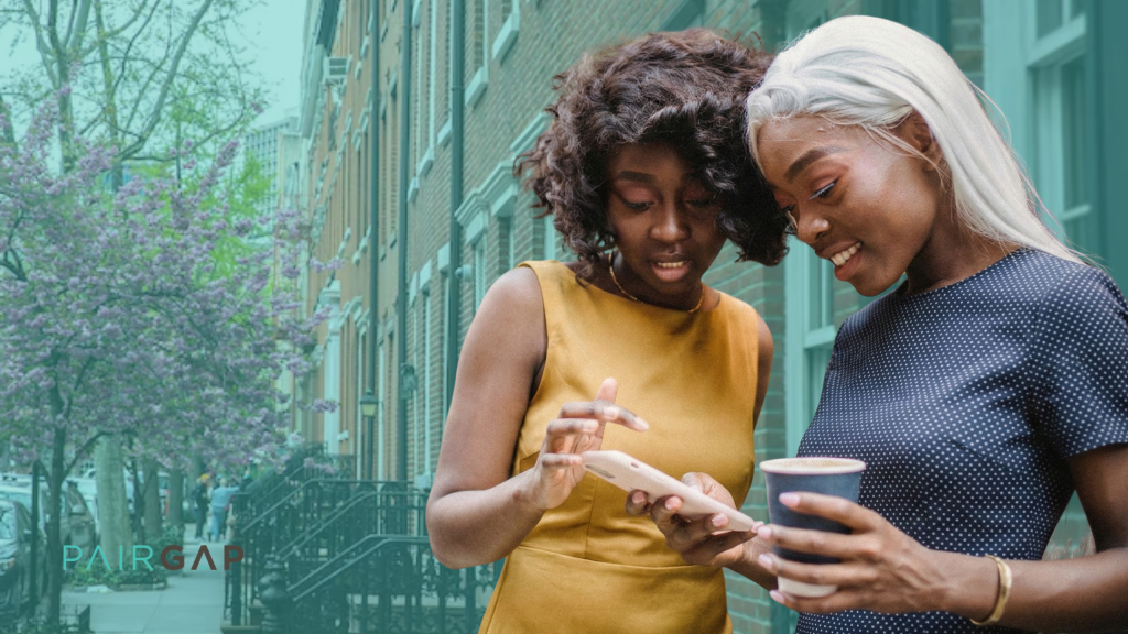 Two women stand on a tree-lined city sidewalk in spring, smiling as they look at a phone together, with the Pairgap logo visible in the lower left corner.